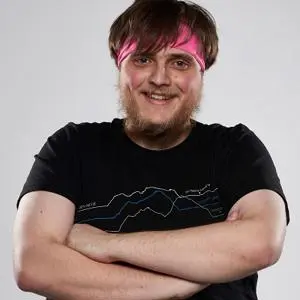 Smiling young man with pink-streaked hair stands with arms folded against a pale background.