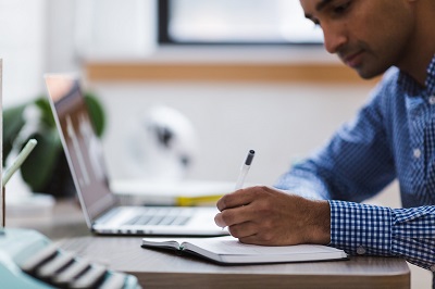 A man writing in a notebook in front of a laptop