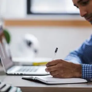 A man writing in a notebook in front of a laptop