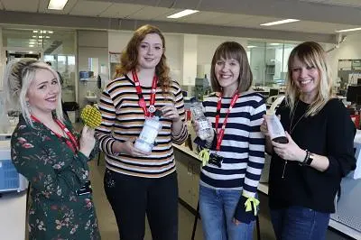 Four women in a laboratory hold sample bottles and protective gloves.