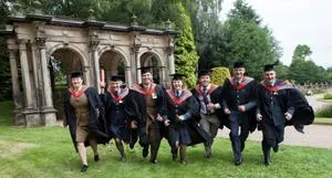 Group of graduates in gowns runs across a lawn in front of a stone monument.