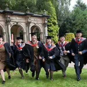 Group of graduates in gowns runs across a lawn in front of a stone monument.