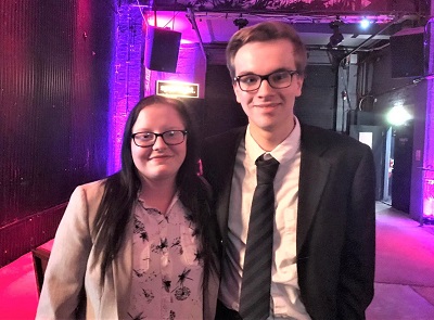 Woman and man pose together at a purple-lit media awards venue.