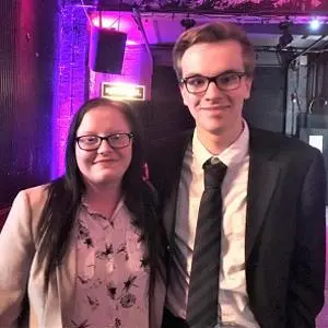 Woman and man pose together at a purple-lit media awards venue.