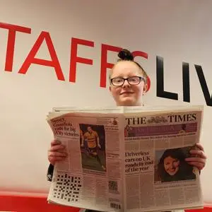 Woman holds an open newspaper beneath large red and black 'STAFFSLIVE' lettering.