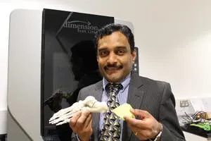 Man in a suit holds a skeletal foot model and a yellow insole beside a display screen.