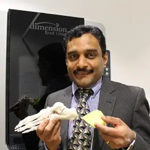 Man in a suit holds a skeletal foot model and a yellow insole beside a display screen.