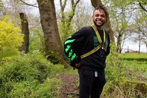 Smiling man in black sportswear and medals stands among trees in a park.