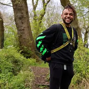 Smiling man in black sportswear and medals stands among trees in a park.