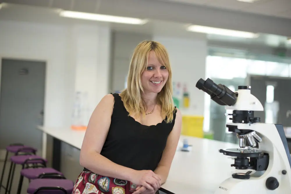 Smiling blonde woman stands beside a microscope in a laboratory.