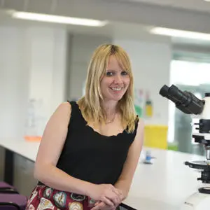 Smiling blonde woman stands beside a microscope in a laboratory.