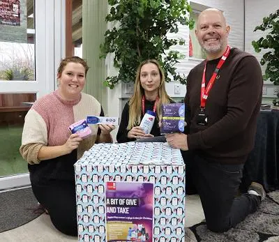 Two people kneel beside a large donation box while holding packaged hygiene products.