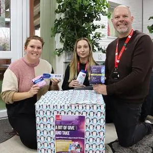 Two people kneel beside a large donation box while holding packaged hygiene products.