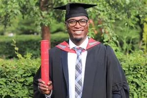 Smiling graduate in cap and gown holds a red diploma tube outdoors.