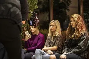 Three women sit on a dimly lit film set while a camera operator works in the foreground.