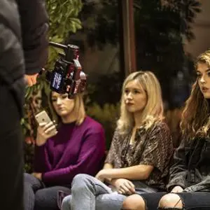 Three women sit on a dimly lit film set while a camera operator works in the foreground.