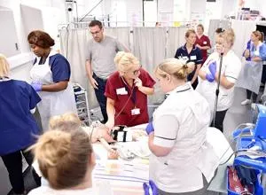 Nursing students and tutors gather around a simulated patient in a bright clinical skills lab.