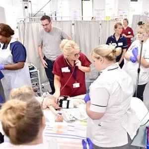 Nursing students and tutors gather around a simulated patient in a bright clinical skills lab.