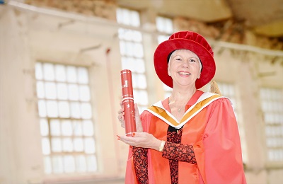 Woman in red academic robes holds a diploma inside a bright industrial-style building.