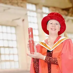 Woman in red academic robes holds a diploma inside a bright industrial-style building.