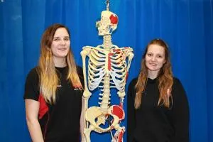 Two smiling women stand on either side of a human skeleton model.
