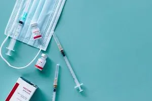 Syringes, surgical masks and vaccine vials arranged on a pale blue surface.