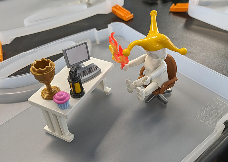 A Playmobil figure sat at a desk holding flames and wearing a jester's hat