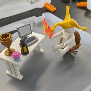 A Playmobil figure sat at a desk holding flames and wearing a jester's hat