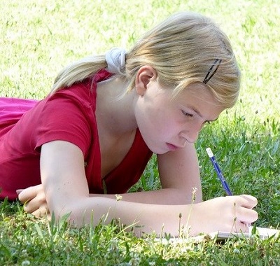 Young girl lies on grass and writes carefully in a notebook.