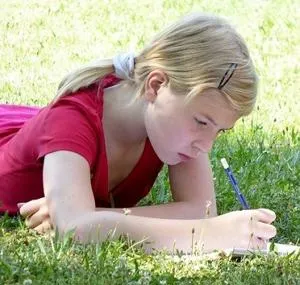 Young girl lies on grass and writes carefully in a notebook.