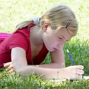 Young girl lies on grass and writes carefully in a notebook.