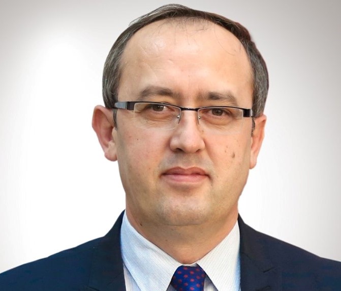 Headshot of a man wearing glasses, a dark suit and patterned tie against a white background.
