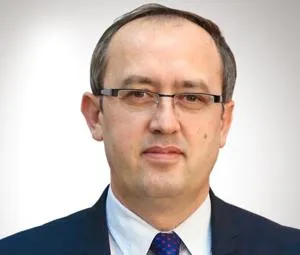 Headshot of a man wearing glasses, a dark suit and patterned tie against a white background.