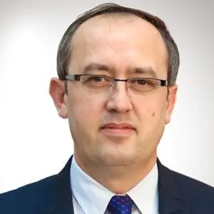 Headshot of a man wearing glasses, a dark suit and patterned tie against a white background.
