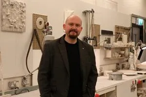 Bald, bearded man in a dark jacket stands inside a ceramics workshop.