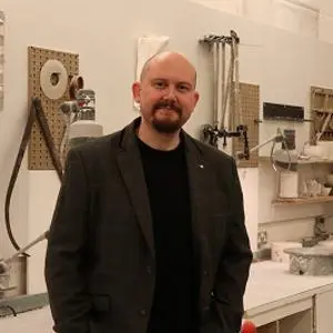 Bald, bearded man in a dark jacket stands inside a ceramics workshop.