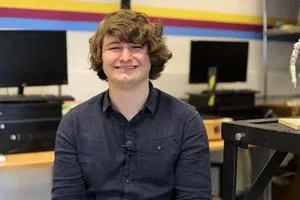 Smiling young man sits in a computer room with monitors and equipment behind him.