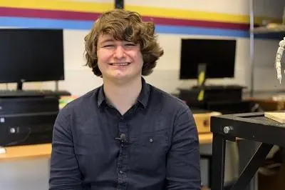 Smiling young man sits in a computer room with monitors and equipment behind him.