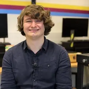 Smiling young man sits in a computer room with monitors and equipment behind him.