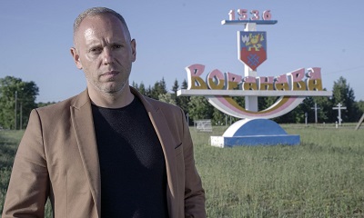 Man in a dark jacket stands in front of a large town sign in a rural landscape.