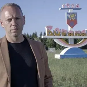 Man in a dark jacket stands in front of a large town sign in a rural landscape.