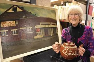 Smiling woman holds a painting of the Victoria Theatre and an 'A Vic Appeal' booklet.