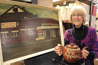 Smiling woman holds a painting of the Victoria Theatre and an 'A Vic Appeal' booklet.