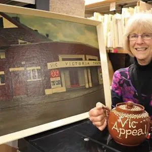 Smiling woman holds a painting of the Victoria Theatre and an 'A Vic Appeal' booklet.