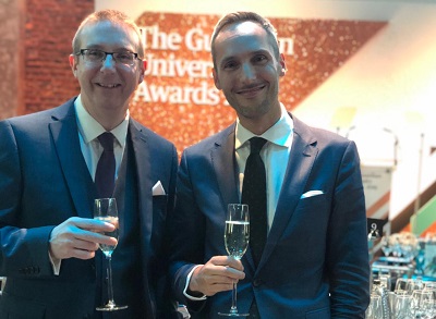 Two men in suits hold champagne glasses at a university awards event.