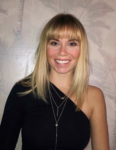 Smiling blonde woman in a black one-shoulder top poses against a pale patterned wall.