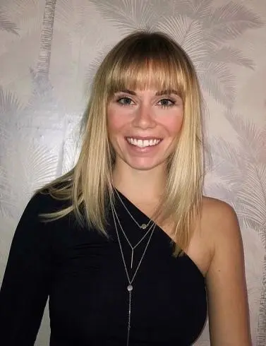 Smiling blonde woman in a black one-shoulder top poses against a pale patterned wall.