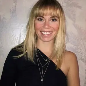 Smiling blonde woman in a black one-shoulder top poses against a pale patterned wall.