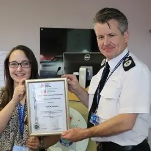Smiling woman and uniformed police officer hold a framed certificate in an office.