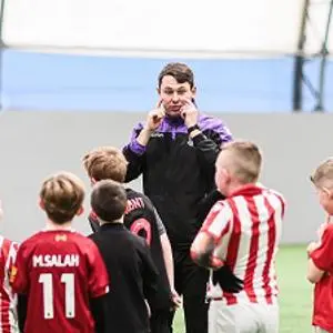 Football coach speaks to a group of children wearing red, white and green kits.